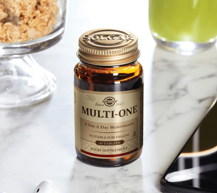 Solgar Multi-One multivitamin bottle on a marble surface with a glass of juice and cereal in the background.
